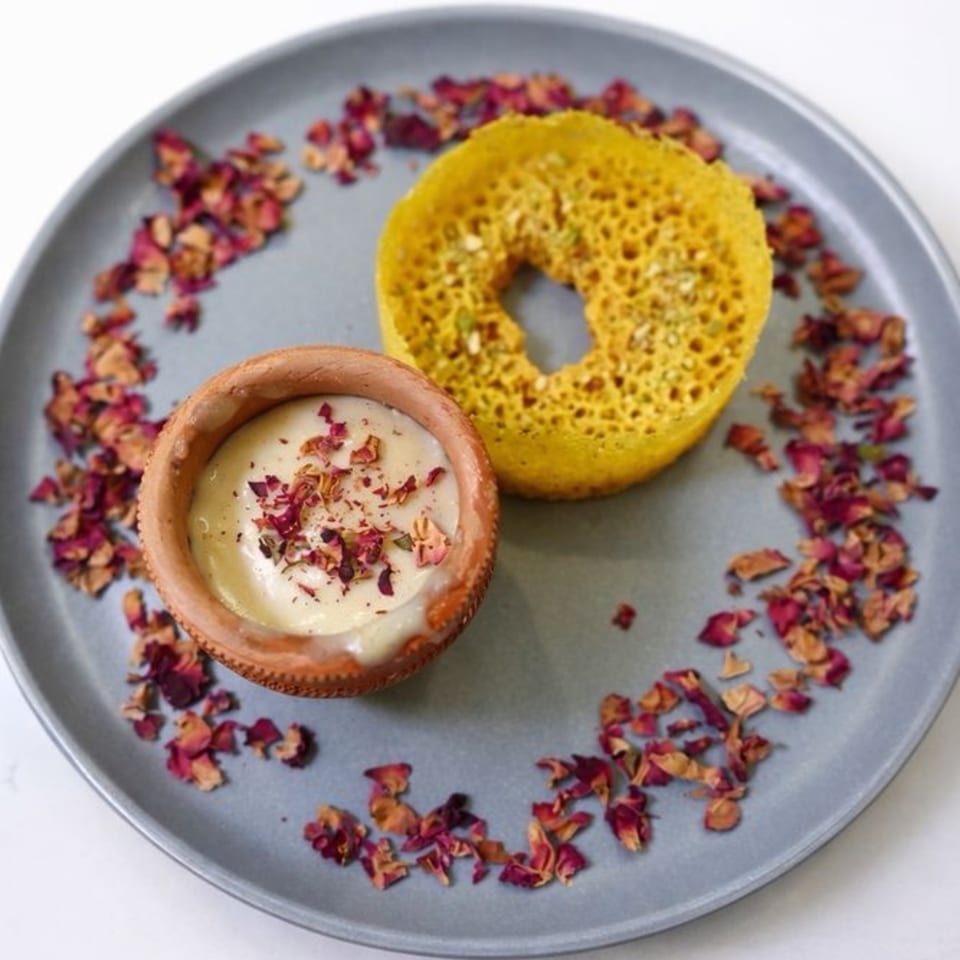 A yellow Indian dessert with a hole in the middle, next to a small clay pot of cream, garnished with rose petals.