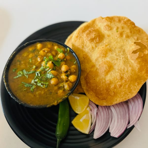 A black plate with chole bhature, garnished with cilantro, lemon wedges, red onion, and a green chili.
