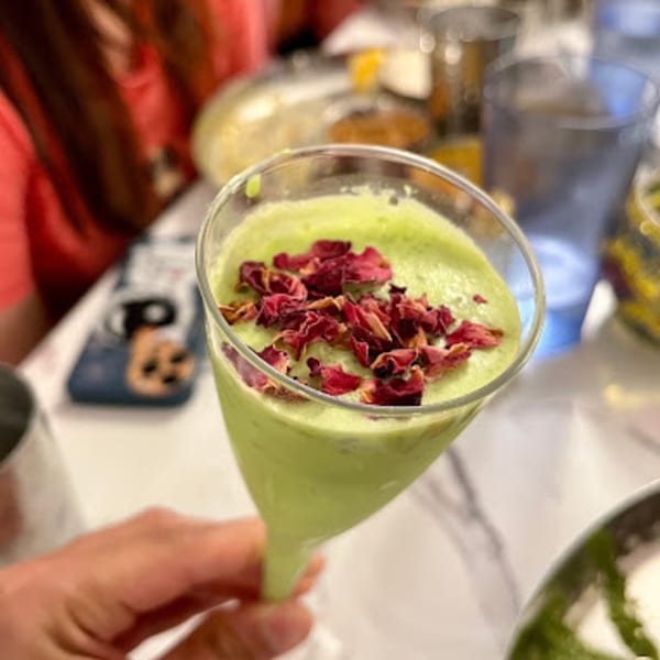 A hand holds a green Indian drink topped with red rose petals in a martini glass.