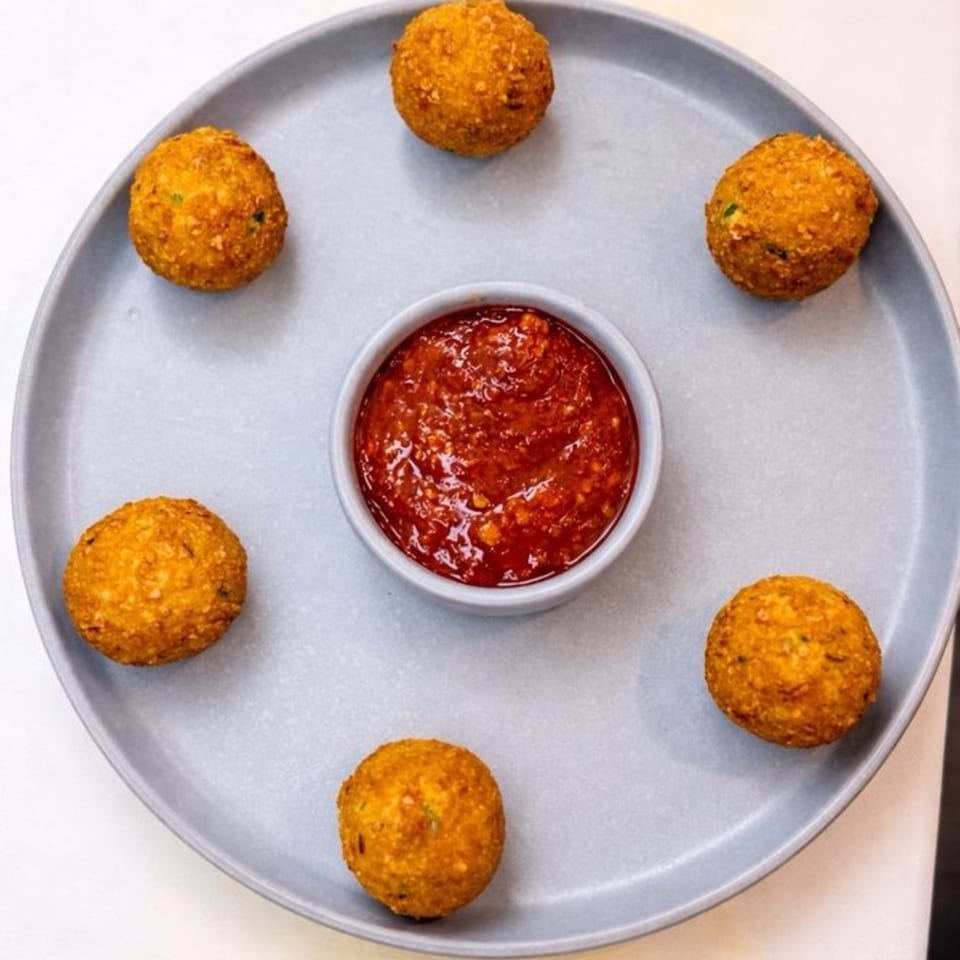 Six golden-brown fried Indian appetizers arranged around a small bowl of red chili sauce on a gray plate.