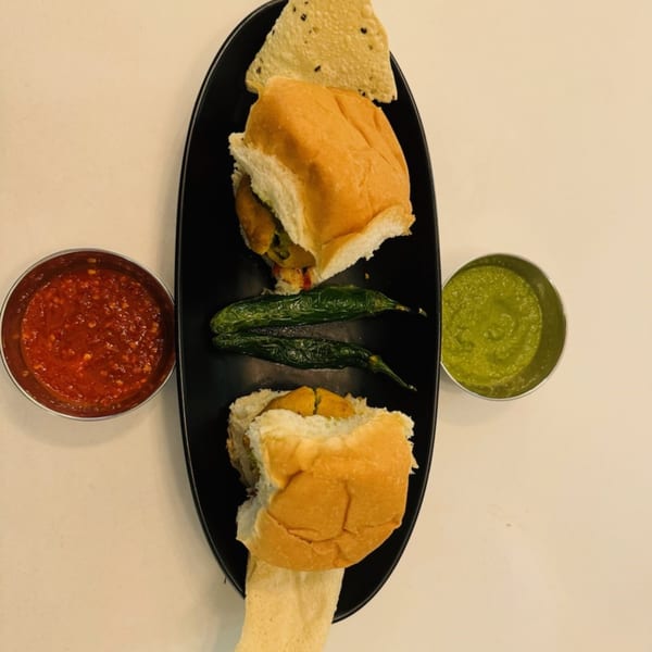 Two vada pavs with fried green chilies, red chutney, and green chutney on a black plate.