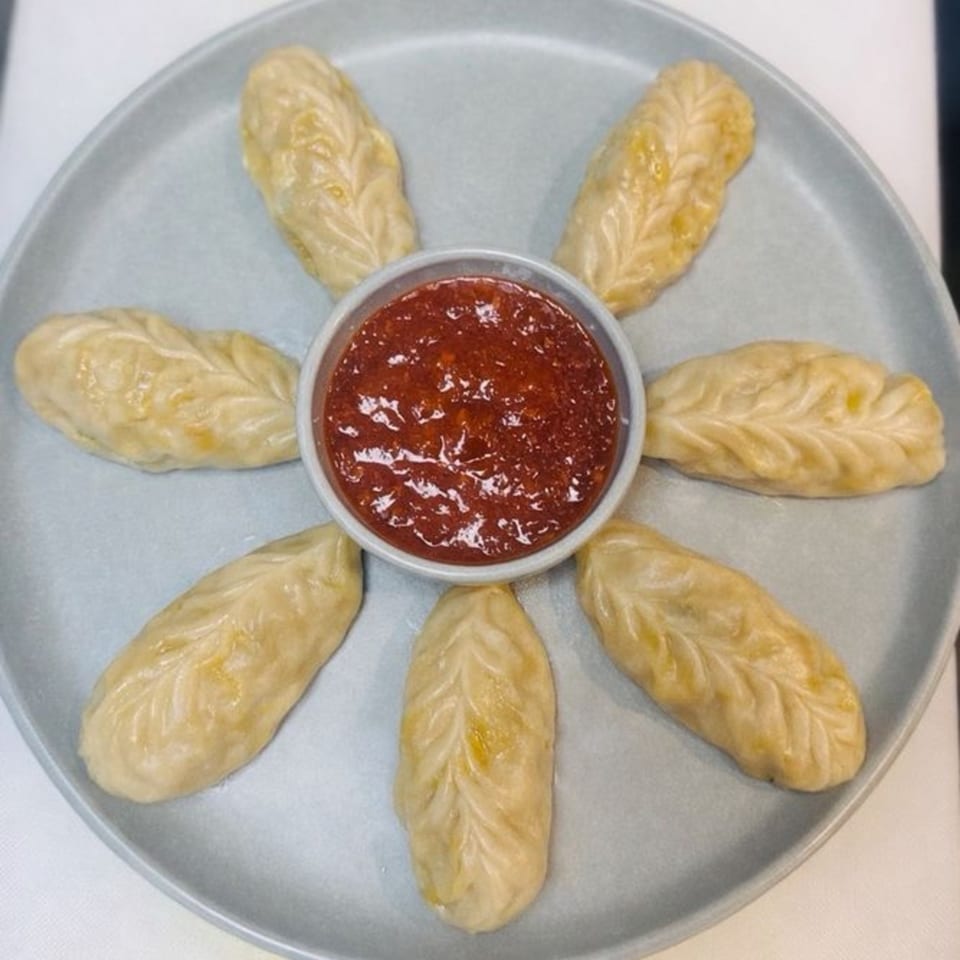 Seven leaf-shaped momos arranged around a small bowl of red chili sauce on a grey plate.